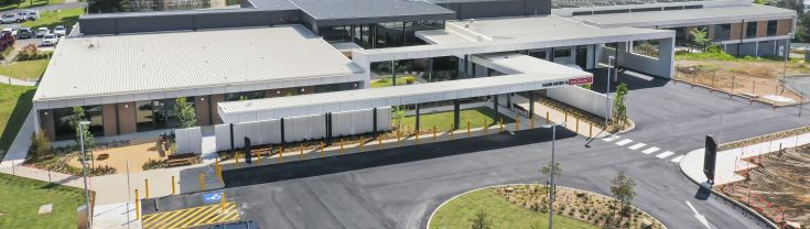 An aerial view of Tumut Health Service. A circular driveway is shown in the bottom-right of the image. Some parking spots, including an accessible parking space sits to the bottom right of the image. The main and emergency entry points is marked on the building. Residences and green spaces spread out behind the building. 