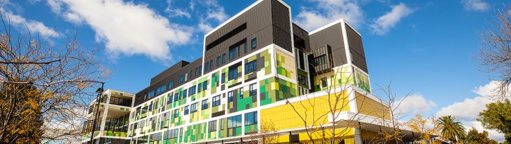 Exterior view of the Wagga Wagga Health Service building. The building features shades of green and yellow clad ties along the bottom and middle and black cladding at the top. A ramp is shown to the far right of the image. Steps and outdoor seating among landscaped green spaces is shown in the foreground of the image. To the left, car parking spaces sit in front of the main entrance. 