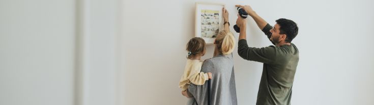 Man using drill while woman holds a picture to a wall while also holding a child.