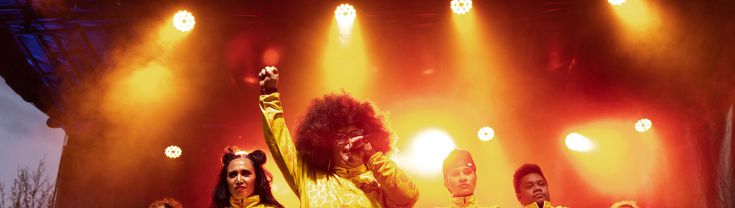 7 people stand with their arms crossed and the leader with her arm in the air and fist closed, holding a microphone and all wearing yellow jumpsuits