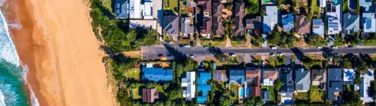 Aerial view of a beachside suburb with roofs, gardens and the sand visible.