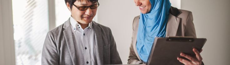 Two people look at a tablet together. One holds a clipboard and wears a bright blue hijab, the other wears a grey suit.