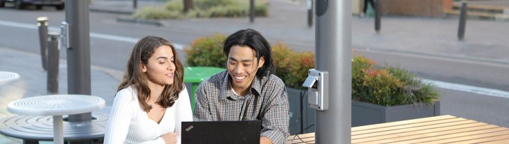 Two people are sitting outside at a ChillOUT Hub in the Georges River Council area, using the provided wi-fi and powerpoints with a laptop. Credit: Georges River Council