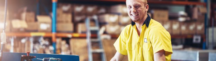 A smiling young man leans on a workbench in a workshop.