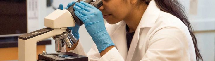 A woman in a white lab coat, looking through a microscope