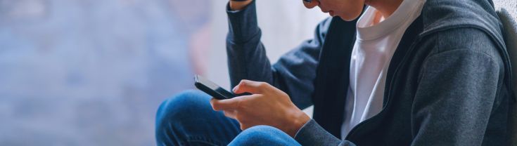 A young man is sitting in a quiet place outside looking stressed with his phone in his hand.