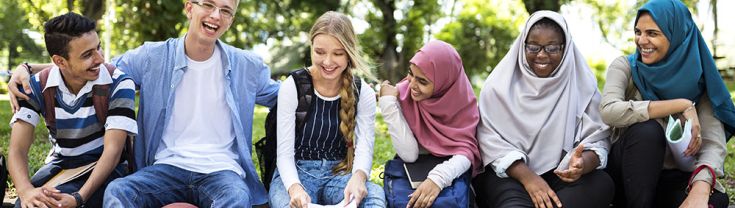 A group of six young people sit on a concrete gutter in a row. All of them are smiling or laughing.There are trees in the background