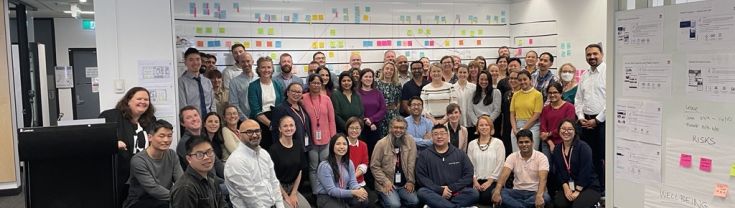 A large group of NSW Digital Channels staff gathers for a group photo during the final 2024 PI (Program Increment) planning session. The room is bright and organised. Office supplies are on a table in the foreground, whilst whiteboards and walls are covered in colourful sticky notes and planning documents in the background. 