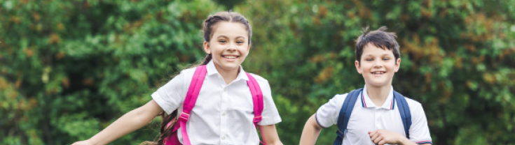 A photograph of two young students, in school uniforms with backpacks on, running outdoors