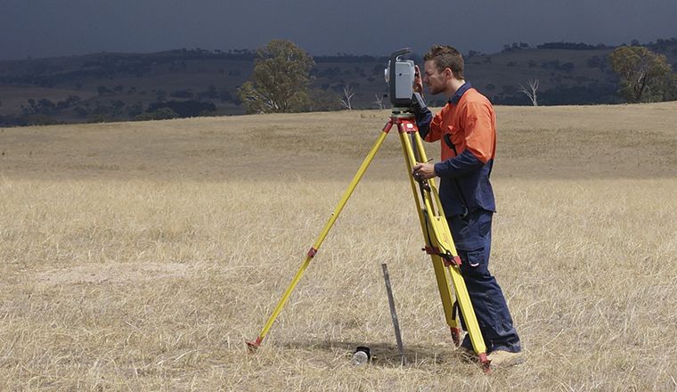 Surveyor with yellow tripod and measuring device in a dry field with dark cloud