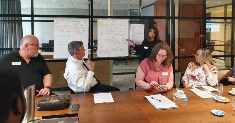 Three men and two women sit at a table watching another woman who stands and points at the group notes on a wall