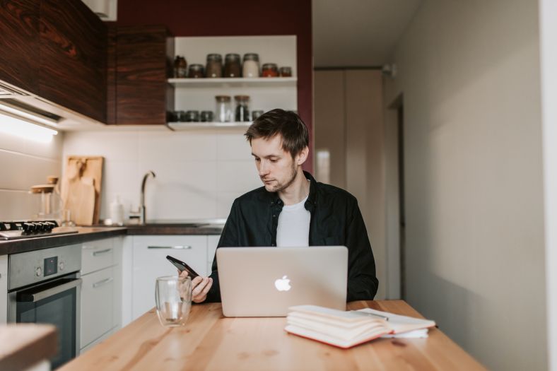 man looks at mobile phone while sitting in front of laptop