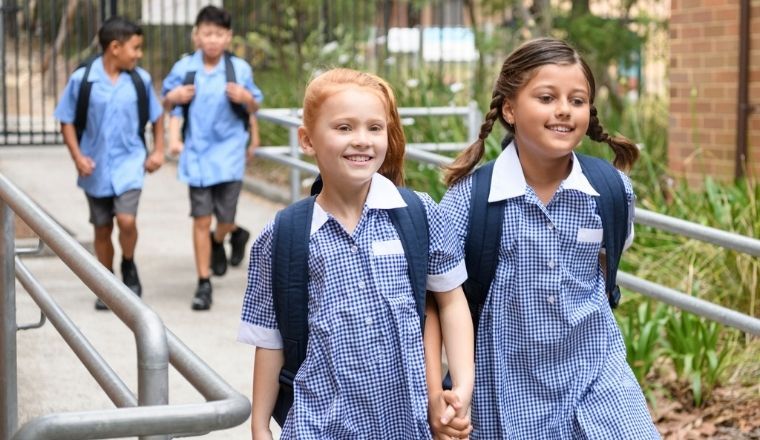 Two school girls holding hands on way to school with a group of boys in the background.