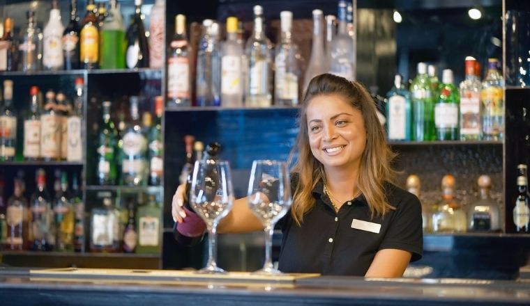 A bartender woman standing and smiling, pouring red wine into a glass from a bottle. Shelves with bottles of alcohol in the background.