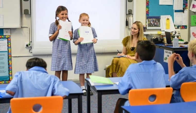 Two school girls presenting their work to their class with their teacher is watching.