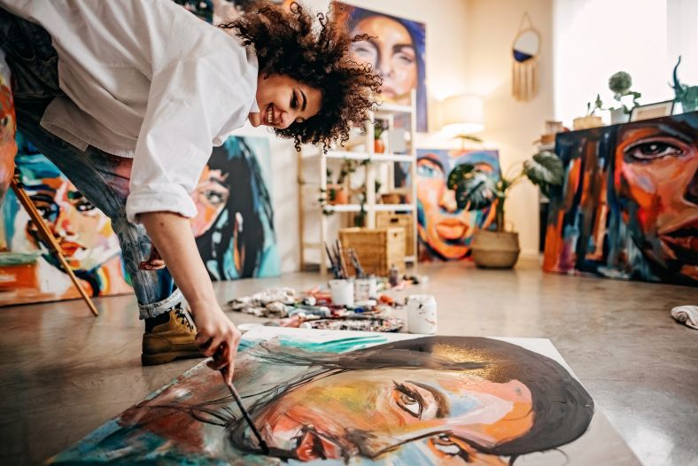 A woman visual artist is smiling as she paints on a colourful canvas spread out across the floor.