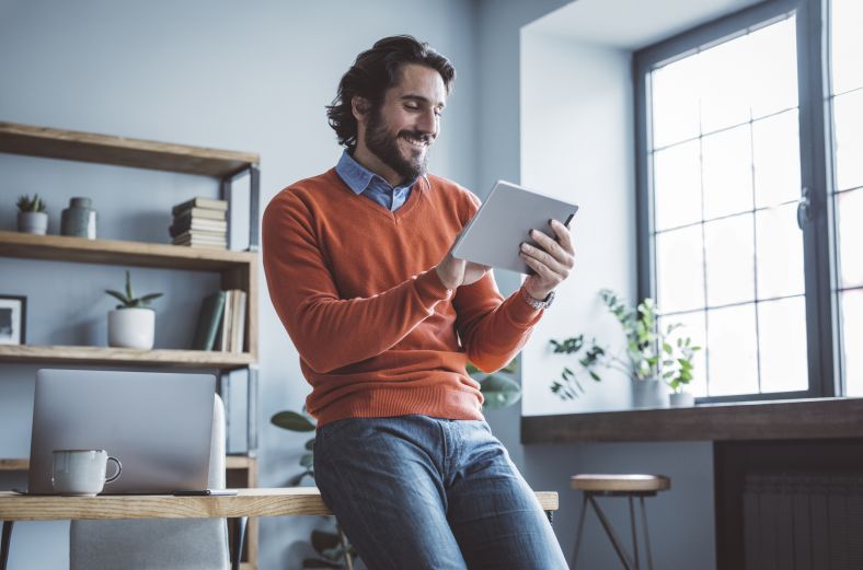 Man, smiling, sitting against desk, looking at device