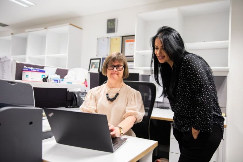 A disabled worker in front of a computer with a supportive boss showing her the way