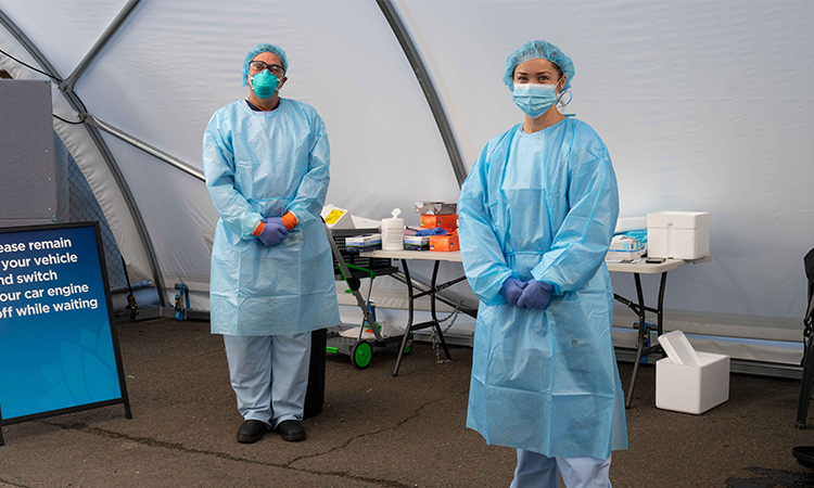 Two people stand, dressed head to toe in blue protective covering and masks