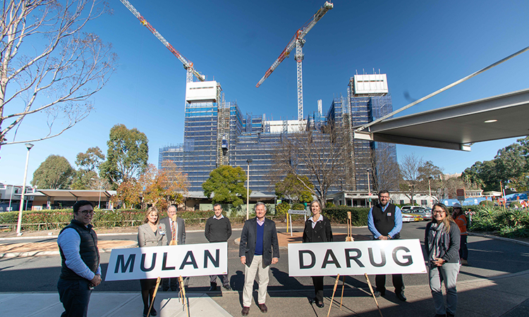 Group of 8 people stand with signs saying Darug and Mulan, with hospital and cranes behind