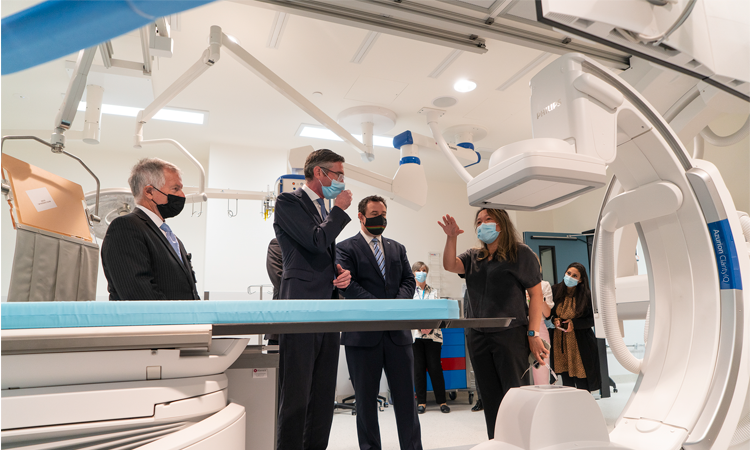 Men in suits and face masks listen to woman in Health uniform talk about a machine they are standing next to