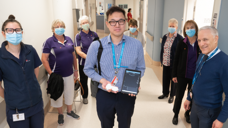 Group of men and women in various health uniforms and masks stand in a corridor, with a man in front holding an ultrasound device and tablet 