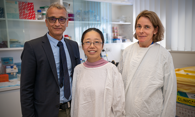 Man in suit stands beside 2 women in white lab coats in a lab