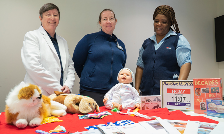 Three people stand smiling behind a table that has soft toys, a doll and some board games on it