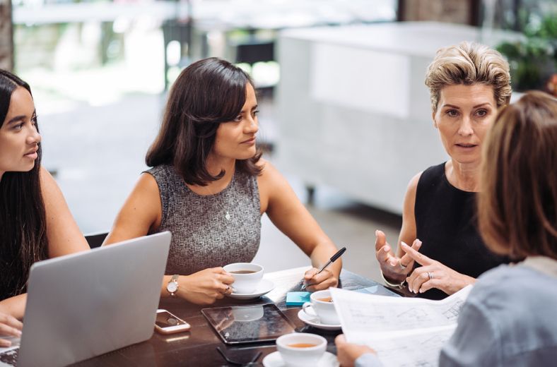 Four women around a table with laptops, paper documents and coffee discussing their strata scheme.