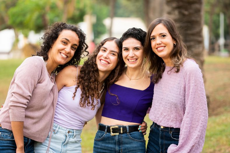 Four women standing together smining at the camera in a park. It is a sunny day and all women are wearing various shades of purple.