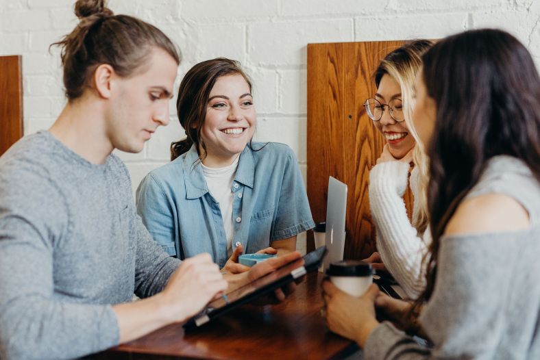 Four young people stand around a wooden tablet holding tablets, computers and coffee. They talk and smile at each other