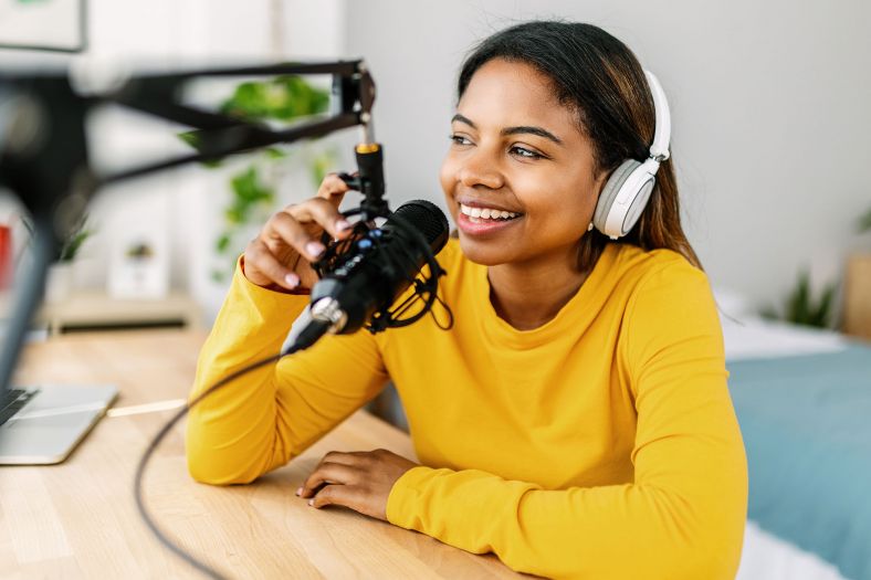 A young woman in a yellow long sleeve top wears white headphones and speaks into a suspended black microphone