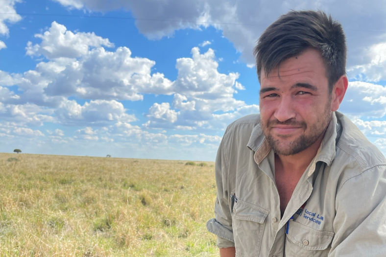 A person with short dark hair smiles while wearing a khaki coloured shirt and kneels in a paddock, with a blue sky and clouds overhead.