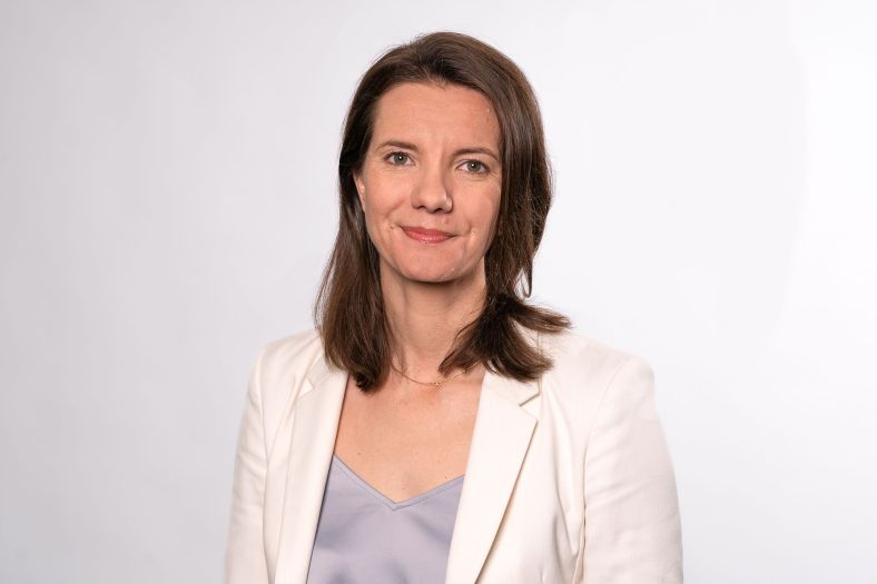 A person with should length brown hair smiles at the camera. She wears a white suit jacket with a gray blouse