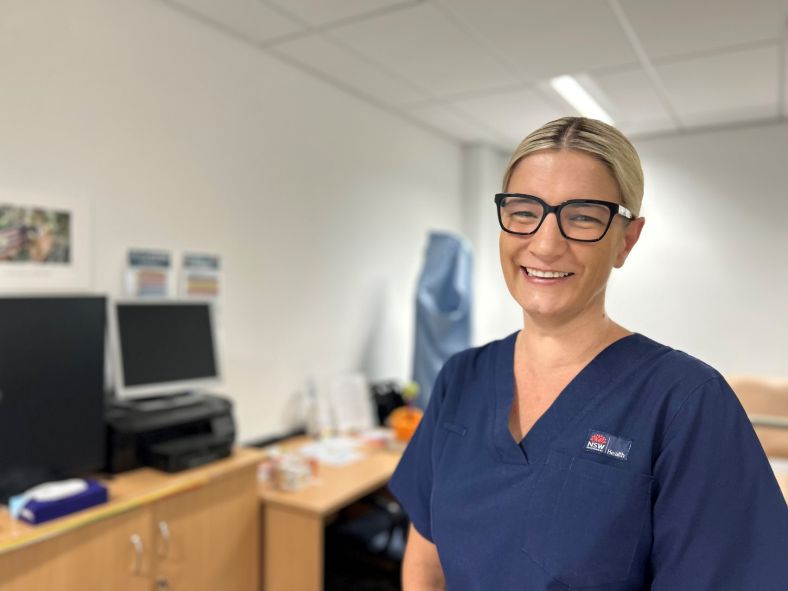 Image of Kate Warren, a Movement Disorder Clinical Nurse Specialist at MLHD. She is wearing a blue nurse uniform and standing in front of an MLHD facility workstation.