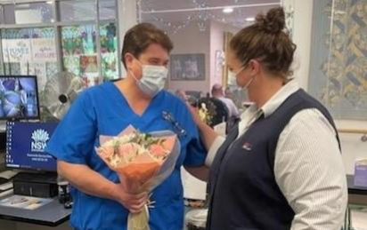 L-R Sharon Longmore wears a blue nurses uniform and face mask, she accepts a flower bouquet from Nurse Unit Manager Carly Silberberg who is wearing a white shirt and black sleeveless sweater and a surgical facemask. They are standing inside Junee hospital.