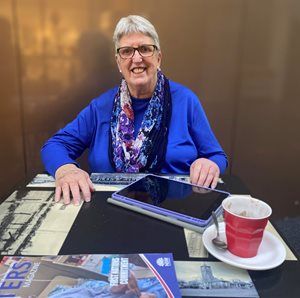 Image of Therese Bartley. She is smiling and sitting at a table wearing a blue stop and multi-coloured scarf. A coffee cup and a tablet is on the table.