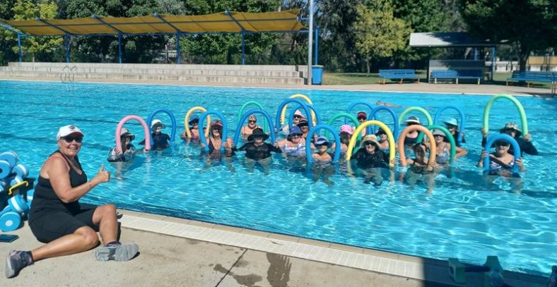 Image of an Aqua Fit class in Young, NSW. Participants are in a swimming pool and the instructor is sitting on the pool's edge showing a thumbs up sign with her right hand.