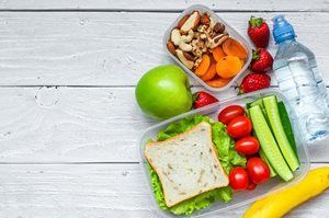 An image of a healthy school lunch placed toward the right of the image. The school lunch consists of a green apple, some nuts and dried fruits in a small container, a larger box with a sandwich and small serve of cucumber, strawberrries and cherry tomatoes, a banana and a bottle of water.