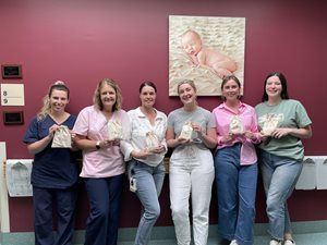Image of midwives from Young, Ashleigh, Shipp, and Kirrilee standing against maroon-red wall with a painting toward the right of the image. They are all holding gift packs to be given to new mothers