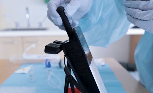 Image shows a gloved hand using a testing device in a dental setting. The device has a black probe and a flat screen that is used to test dental unit waterlines for biohazards.