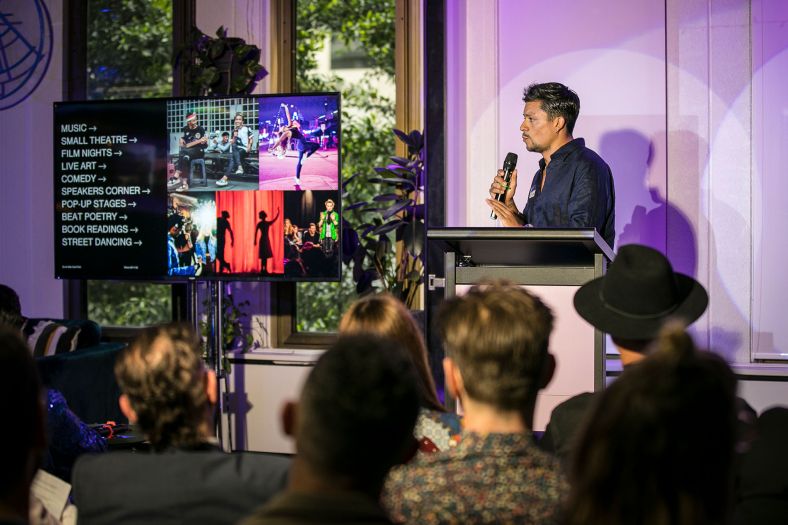 Person speaking on stage with a microphone and looking at a screen