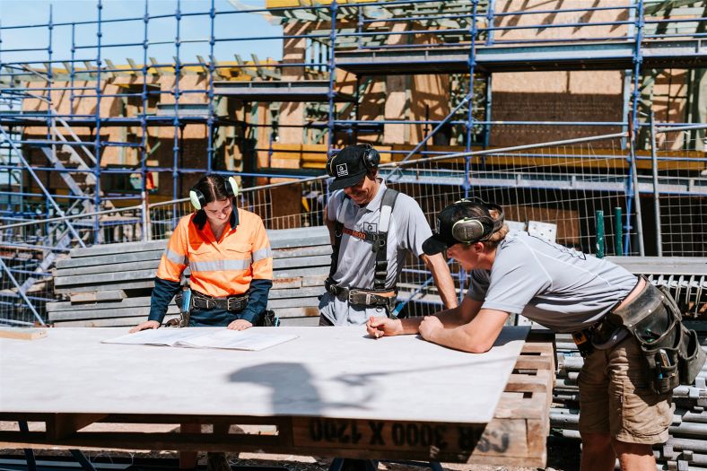 Three workers are standing on a construction site laughing as they look over plans.