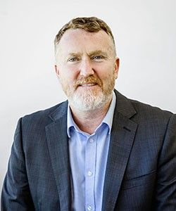 A man with light brown and gray hair with the moustache and beard smiles at the camera. He wears a light blue shirt and dark gray suit jacket