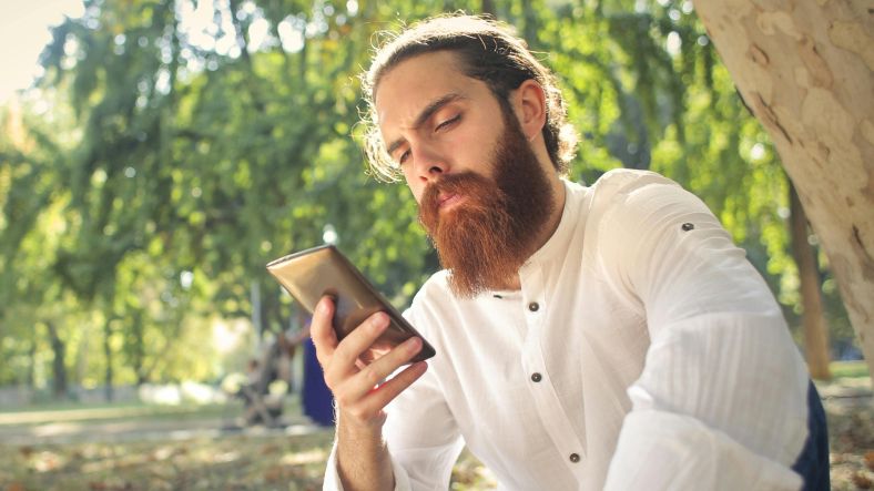 A man sat under a tree holding a mobile phone.