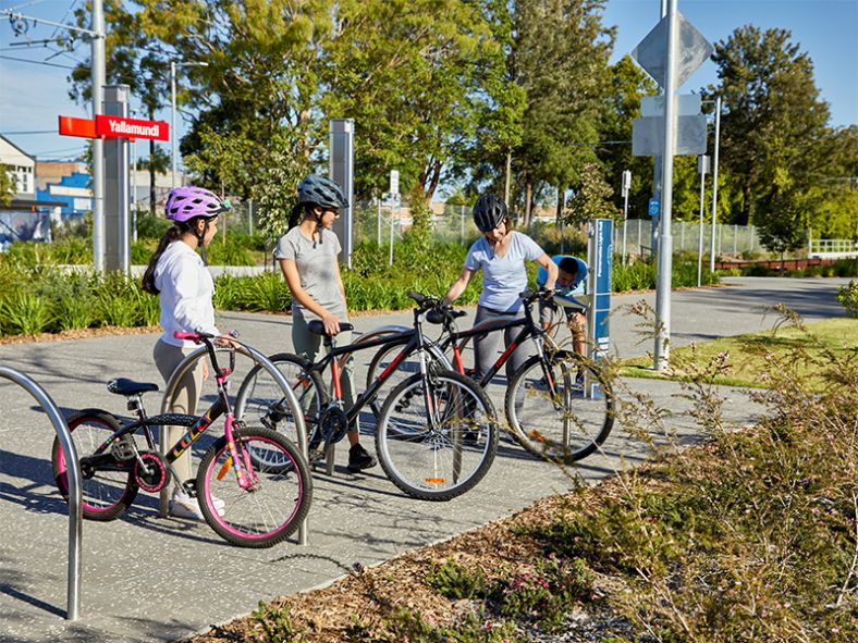Three people wearing helmets with their bikes next to Yallamundi Light Rail station.