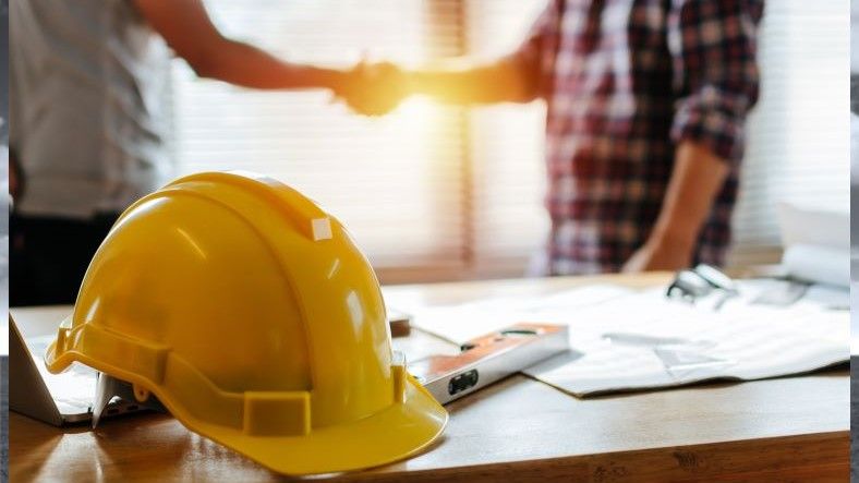 Yellow hard hat on table with torsos only showing of people shaking hands behind.