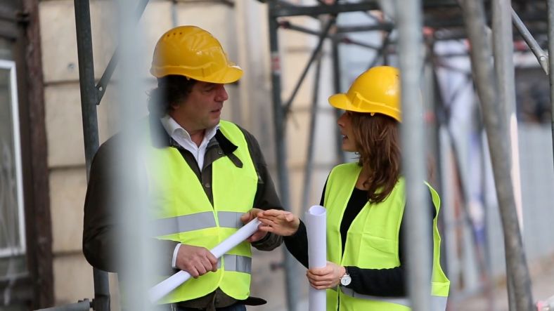 Two people in scaffolding talking, both wearing yellow hard hats and hi-vis vests.