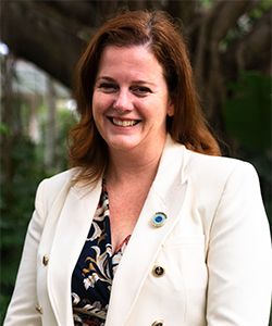 Smiling woman with dark hair in black patterned top and white jacket in a background of greenery