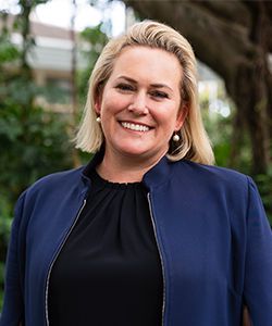 Smiling woman with blond hair in black dress and dark blue jacket in a background of greenery